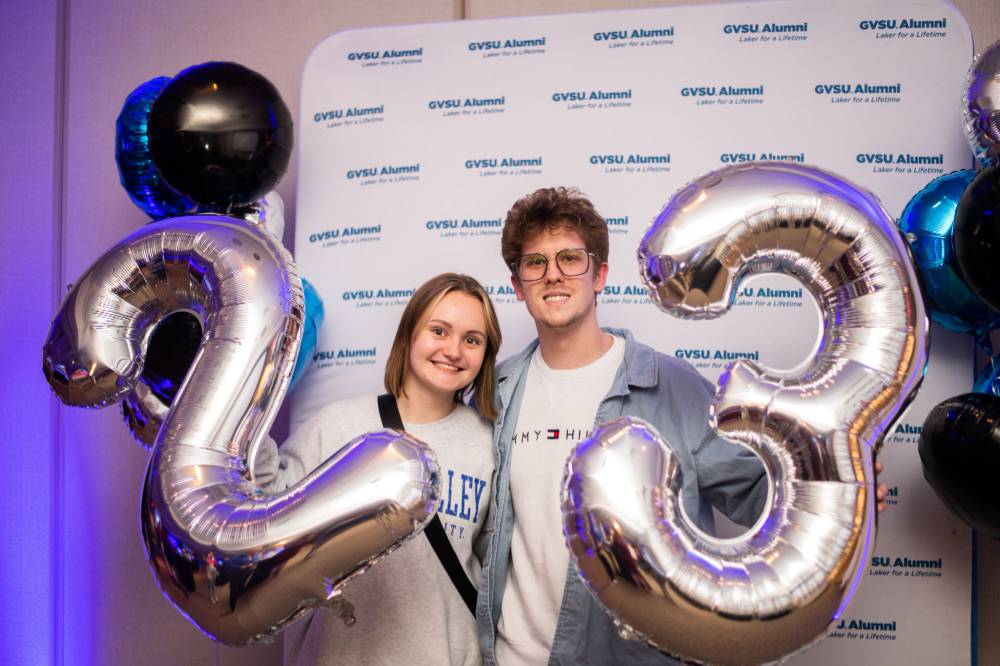 Two grads pose for a photo with the GVSU alumni backdrop holding 2023 balloons
